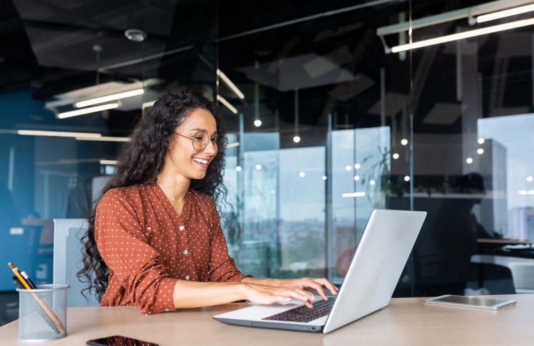 Happy and smiling hispanic businesswoman typing on laptop, office worker with curly hair and glasses happy with achievement results, at work inside office building.